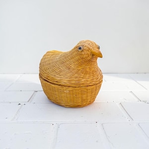 May include: A woven bird-shaped basket with a lid. The basket is made of light brown wicker and features a beak and eyes, giving it a whimsical appearance. The bird-shaped basket is a decorative storage item.