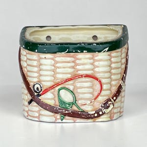 Op de afbeelding: Een keramische plantenbak in de vorm van een mand met een groene rand en bruine banddetails. De mand heeft een hengel- en netontwerp in rood en groen. De plantenbak heeft een geweven mandpatroon.