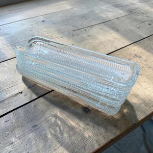 May include: A stack of clear glass rectangular serving dishes. Each dish has a textured, ribbed pattern and a small handle on one end. The dishes are stacked on a weathered wooden surface.