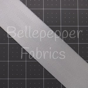 May include: A close-up of a gray woven webbing ribbon. The ribbon is on a black grid background.