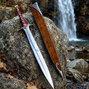 May include: A silver metal sword with a red wrapped handle and intricate silver crossguard, accompanied by a brown wooden scabbard featuring silver metal accents at the top and bottom, resting against a large gray rock surface.