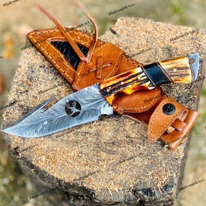 Peut inclure: Un couteau de chasse avec une lame à motifs et un manche en os et en bois. Le couteau est dans un étui en cuir marron avec une fermeture à bouton-pression. La lame a une silhouette de cerf découpée.