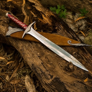 May include: A silver-toned metal sword with a pointed blade and an ornate crossguard. The handle features a deep red grip with silver filigree patterns. The sword rests partially inside a brown suede-like scabbard with silver metal accents.