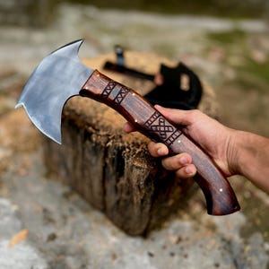 May include: A handcrafted axe with a polished steel blade and a dark wood handle. The handle features intricate carved designs and is held by a hand. The axe is resting on a wooden stump, with another axe visible in the background.