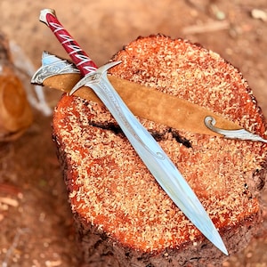 May include: A silver-toned sword with a pointed blade and a crossguard featuring intricate engravings. The handle is deep red with silver accents. A tan-colored scabbard with silver metal detailing at the tip and throat rests beneath the sword.