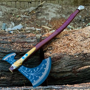 May include: A double-bitted axe featuring a dark blue head with white etched runic patterns and a gold-colored metal accent piece with two blue circular inlays. The long handle is wrapped in dark maroon leather with a silver-colored metal pommel.