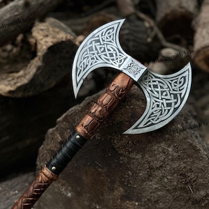 May include: A double-bladed axe with a silver head featuring intricate Celtic knot designs. The wooden handle is carved with detailed patterns and wrapped with black leather near the center. The axe is displayed on a natural wood background.