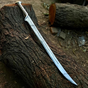 May include: A silver-toned metal sword featuring an elongated, curved blade with intricate, etched vine-like patterns running along the center and handle. The hilt has a rounded pommel and a pointed guard, with the text BladesmithArts visible.