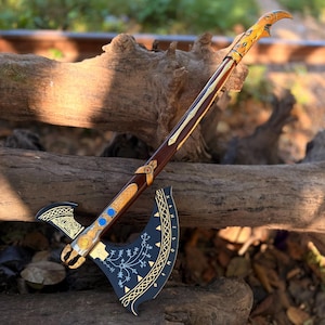 Puede incluir: Un hacha decorativa detallada con un mango de madera marrón oscuro y una hoja negra. El cabezal del hacha presenta detalles dorados y azules, con patrones intrincados. El mango tiene detalles dorados y marrones. El hacha se exhibe sobre un tronco de madera desgastado.