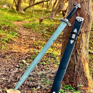 Puede incluir: Una espada larga y plateada con una hoja estampada y una empuñadura adornada descansa contra un árbol, junto a una vaina negra. El mango de la espada es negro y la vaina tiene detalles plateados. La escena está ambientada al aire libre, con un fondo de bosque.