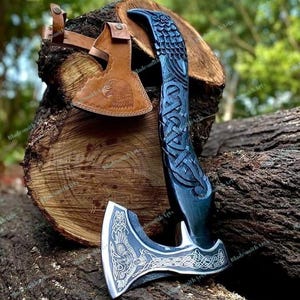 Pode incluir: Um machado de mão com cabo de madeira escura esculpida com nós intrincados e lâmina de metal prateado gravada com um motivo de pássaro. Uma bainha de couro castanho com um design de cabeça de águia em relevo está ao lado da lâmina.