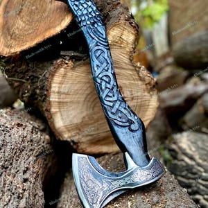 May include: A hand-forged axe with a raven head design and intricate Celtic knotwork carvings. The axe head is silver, and the handle is dark gray. The axe is displayed on a wooden log.