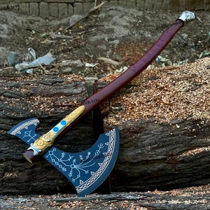 Puede incluir: Un hacha artesanal con una hoja azul oscuro y un mango marrón envuelto en cuero burdeos. La cabeza del hacha presenta elementos decorativos dorados y blancos. El hacha está sobre un tronco de madera, mostrando su detallada artesanía.