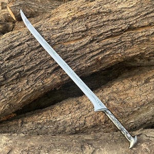 May include: A long, curved sword with a silver blade and an ornate handle. The blade features a detailed etched pattern. The handle is silver with a decorative design. The sword is set against a backdrop of textured brown logs.