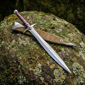 May include: A double-edged dagger with a pointed steel blade featuring etched scrollwork near the hilt. The handle is wrapped in deep purple material with silver accents, and it rests alongside a tan suede scabbard with ornate silver metal fittings.