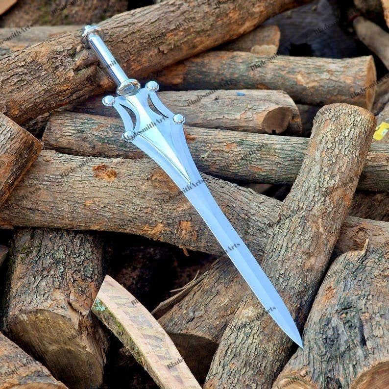 May include: A polished silver-toned metal dagger featuring a pointed blade, a central ridge, and a stylized crossguard with four rounded decorative accents. The handle is cylindrical with a smooth finish, leading to a rounded pommel at the base.
