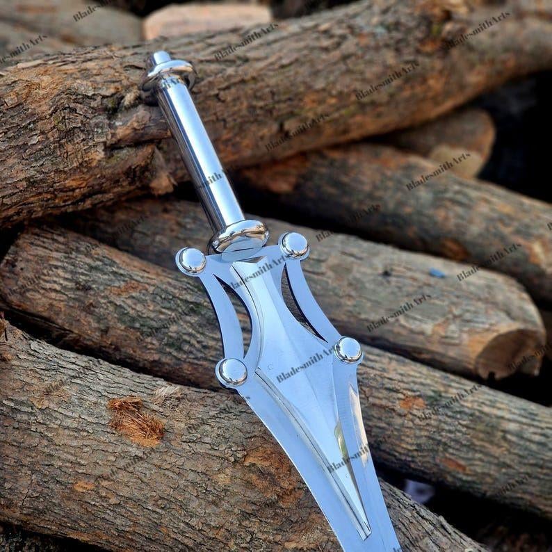 May include: A polished silver-colored metal dagger featuring a cylindrical handle and a wide, pointed blade with symmetrical cutouts and four rounded decorative studs at the crossguard. The blade surface is smooth and reflective.