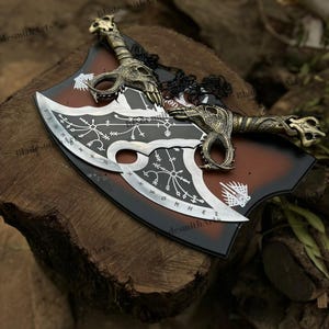 Pu&ograve; includere: Una coppia di lame in metallo argentato con intricati simboli runici e motivi incisi in bianco. Le impugnature presentano dettagliati motivi a teschio color oro e prese testurizzate. Le lame sono montate su una placca in legno marrone e nero.