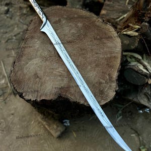 May include: A silver-toned metal sword featuring a curved blade and a handle with intricate, etched vine-like patterns. The blade displays a long, slender design with a pointed tip and decorative engravings running along the center of the steel surface.