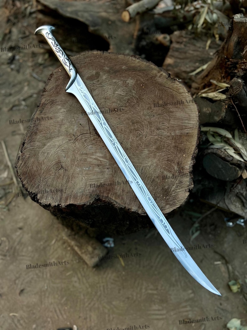 May include: A silver-toned metal sword featuring a curved blade and a handle with intricate, etched vine-like patterns. The blade displays a long, slender design with a pointed tip and decorative engravings running along the center of the steel surface.