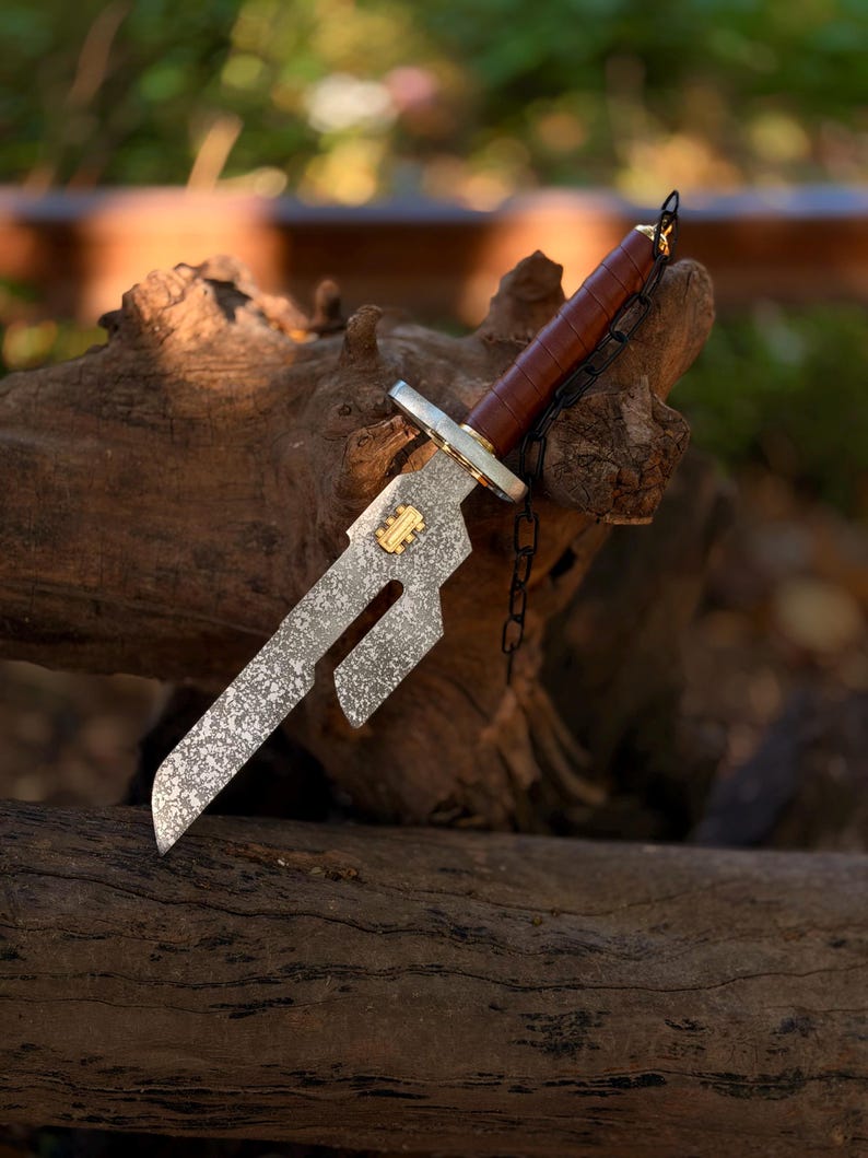 May include: A decorative dagger featuring a mottled grey blade with a distinct cutout section and a small gold-colored rectangular emblem. The handle is wrapped in brown material with a silver-toned crossguard and a thin black chain attached.