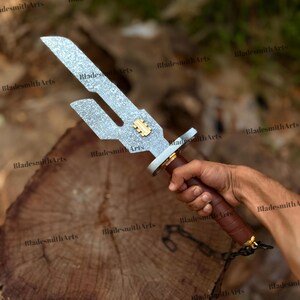 May include: A metal sword with a textured silver blade featuring a distinct cutout notch and a small gold-colored rectangular emblem. The handle is wrapped in brown material with a silver guard and a short black chain attached to the pommel.