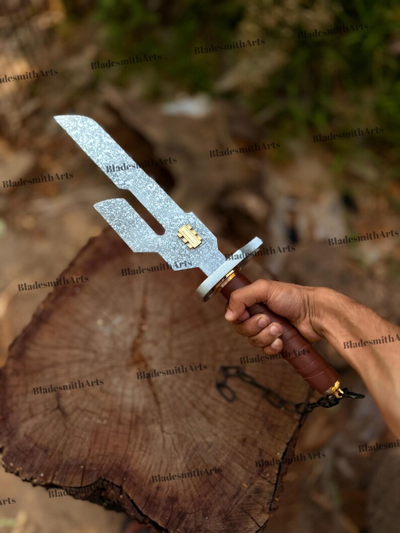 May include: A metal sword with a textured silver blade featuring a distinct cutout notch and a small gold-colored rectangular emblem. The handle is wrapped in brown material with a silver guard and a short black chain attached to the pommel.