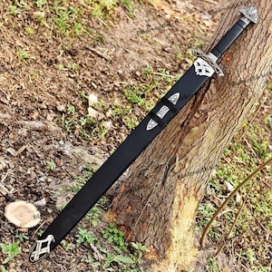 May include: A black sword with a silver handle and accents, resting against a tree trunk. The sword has a long, straight blade and is encased in a black sheath. The handle features intricate silver designs.