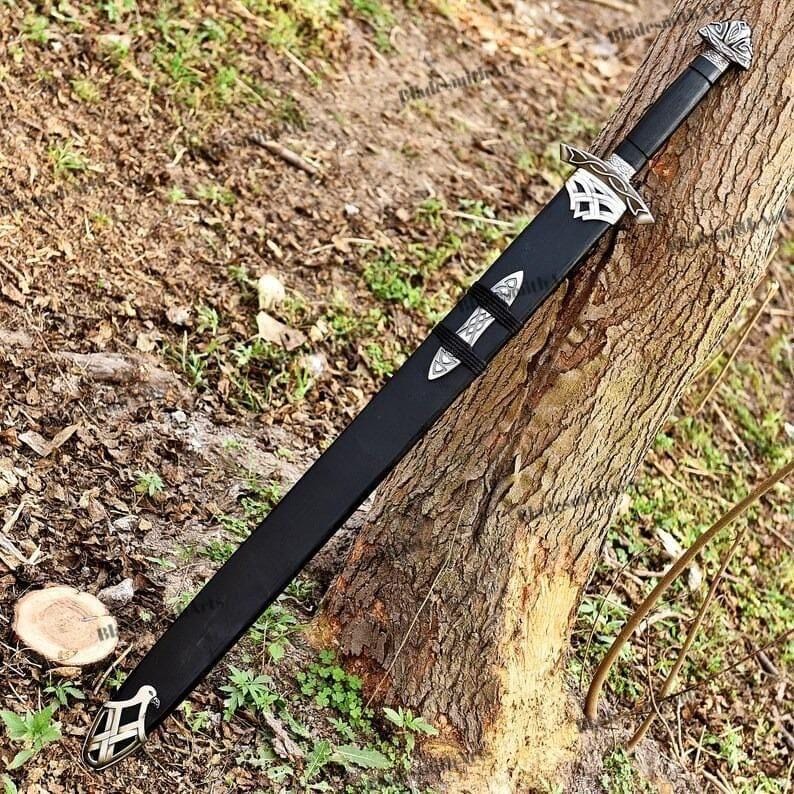 May include: A black sword with a silver handle and accents, resting against a tree trunk. The sword has a long, straight blade and is encased in a black sheath. The handle features intricate silver designs.
