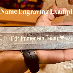 May include: Close-up of a metal object with a patterned design. The text "F&uuml;r immer ein Team" is engraved in white, along with a heart symbol. The words "Name Engraving Example" are also visible.