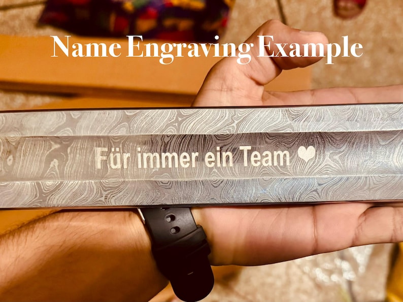 May include: Close-up of a metal object with a patterned design. The text "F&uuml;r immer ein Team" is engraved in white, along with a heart symbol. The words "Name Engraving Example" are also visible.