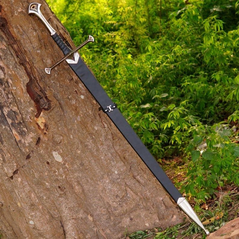 May include: A long sword with a silver-toned pommel and crossguard, featuring a black wrapped grip. The blade is encased in a black scabbard with a silver-toned chape at the tip and a metal fitting near the hilt, resting against a tree trunk.