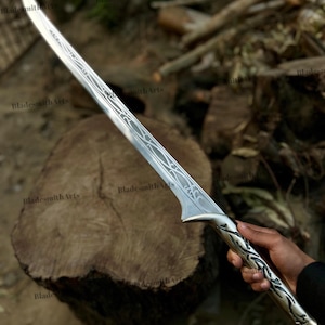 May include: A silver-toned sword featuring an elongated, curved blade with intricate, etched filigree patterns running along the center. The handle is crafted with matching metallic scrollwork designs and a flared pommel at the base of the grip.