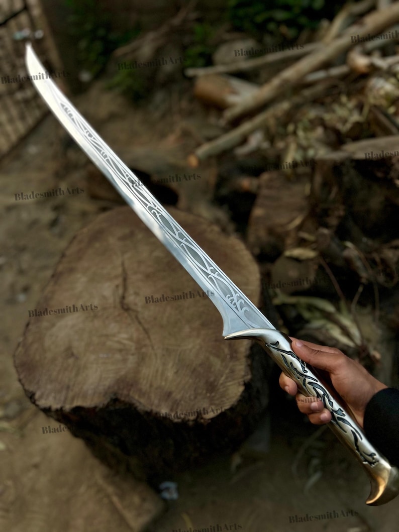 May include: A silver-toned sword featuring an elongated, curved blade with intricate, etched filigree patterns running along the center. The handle is crafted with matching metallic scrollwork designs and a flared pommel at the base of the grip.