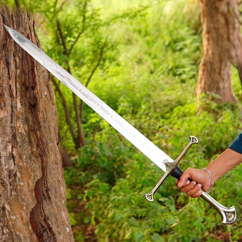 May include: A long, double-edged steel sword with a pointed tip and a metallic crossguard. The blade features an etched inscription near the hilt, which has a dark grip and a flared pommel. The sword is held at an angle against a tree trunk.