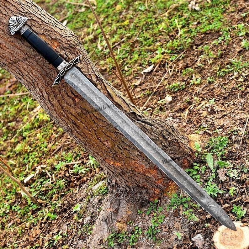 May include: A detailed image of a long, silver sword with a black handle and ornate silver accents. The blade has a textured pattern. The sword is resting on a tree trunk, with green foliage in the background.