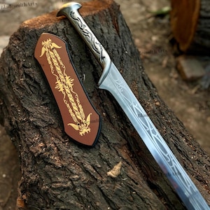 May include: A silver-toned metal sword with an ornate, engraved handle and a long blade featuring intricate, flowing line patterns. Beside the sword lies a brown, shield-shaped plaque decorated with a gold-colored, symmetrical floral design.