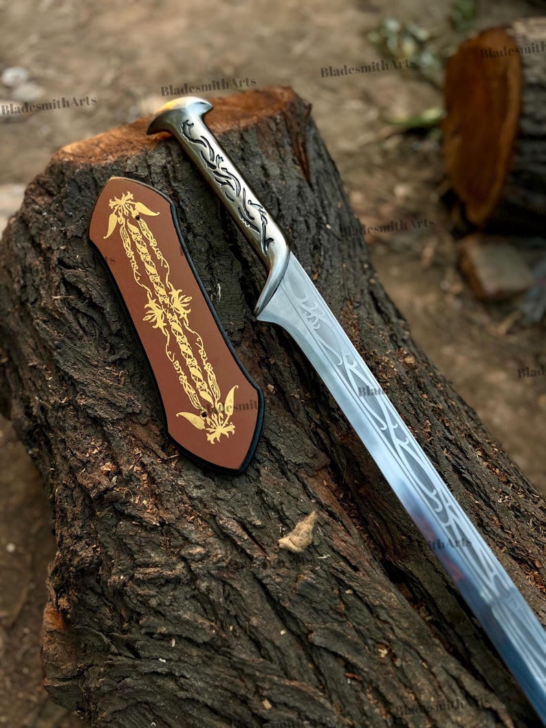 May include: A silver-toned metal sword with an ornate, engraved handle and a long blade featuring intricate, flowing line patterns. Beside the sword lies a brown, shield-shaped plaque decorated with a gold-colored, symmetrical floral design.