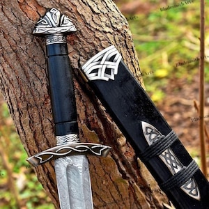 May include: A detailed image of a sword with a black handle and scabbard. The sword features a silver-colored blade with a unique pattern. The handle and scabbard are adorned with intricate silver designs. The sword is resting against a tree trunk.