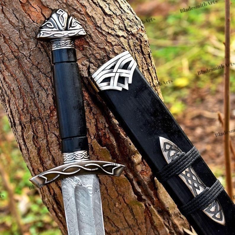 May include: A detailed image of a sword with a black handle and scabbard. The sword features a silver-colored blade with a unique pattern. The handle and scabbard are adorned with intricate silver designs. The sword is resting against a tree trunk.