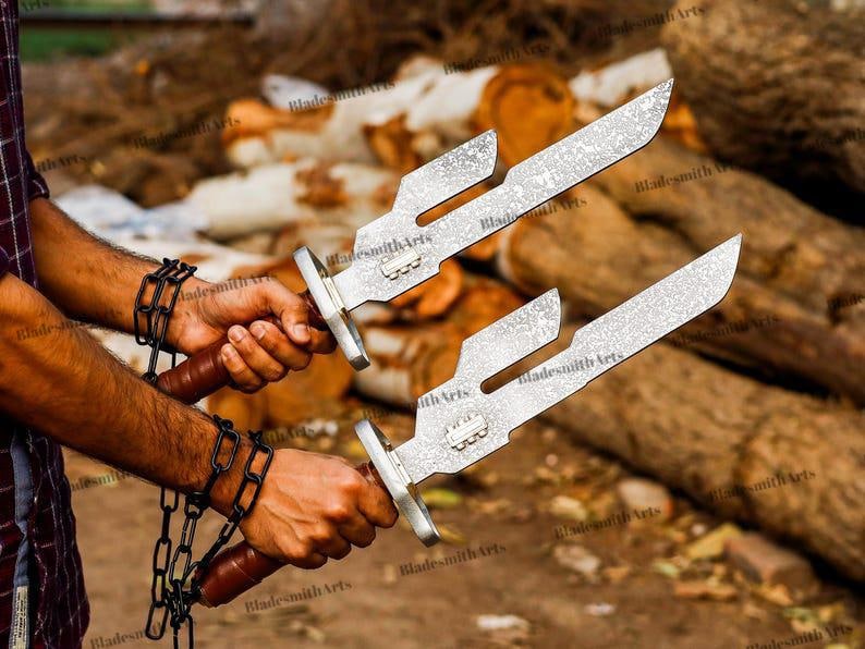 May include: A pair of silver-toned metal blades with a distinct notched design and a textured finish. Each blade features a brown handle and is attached to a black metal chain. The blades have a rectangular metal guard near the handle area.
