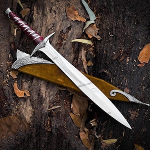 May include: A silver-toned metal dagger with a pointed blade featuring intricate etched patterns. The handle is wrapped in deep red material with silver accents. It is accompanied by a mustard-yellow scabbard with ornate silver metal detailing.