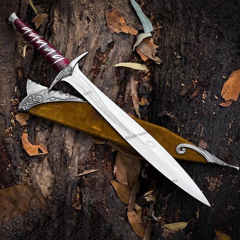 May include: A silver-toned metal dagger with a pointed blade featuring intricate etched patterns. The handle is wrapped in deep red material with silver accents. It is accompanied by a mustard-yellow scabbard with ornate silver metal detailing.