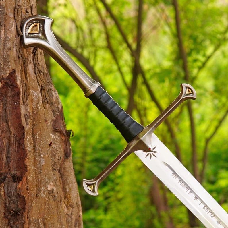 May include: A metal sword featuring a black wrapped grip, a polished silver crossguard with gold-toned accents, and a pommel with a gold inlay. The blade displays an engraved star symbol and runic inscriptions along the central fuller.
