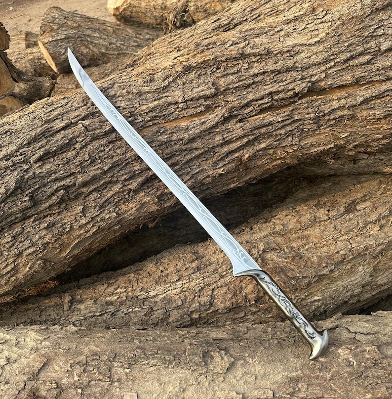 May include: A silver-toned metal sword featuring a long, slender blade with intricate etched vine-like patterns running along its length and a curved handle with matching decorative engravings, presented as a single-edged pointed weapon.