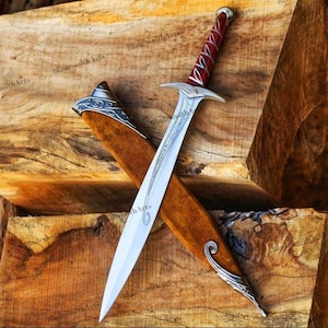 May include: A silver-toned metal sword with a pointed blade featuring etched line details. The handle is wrapped in a deep red material with silver accents. It is positioned diagonally across a brown wooden scabbard with silver metal trim.