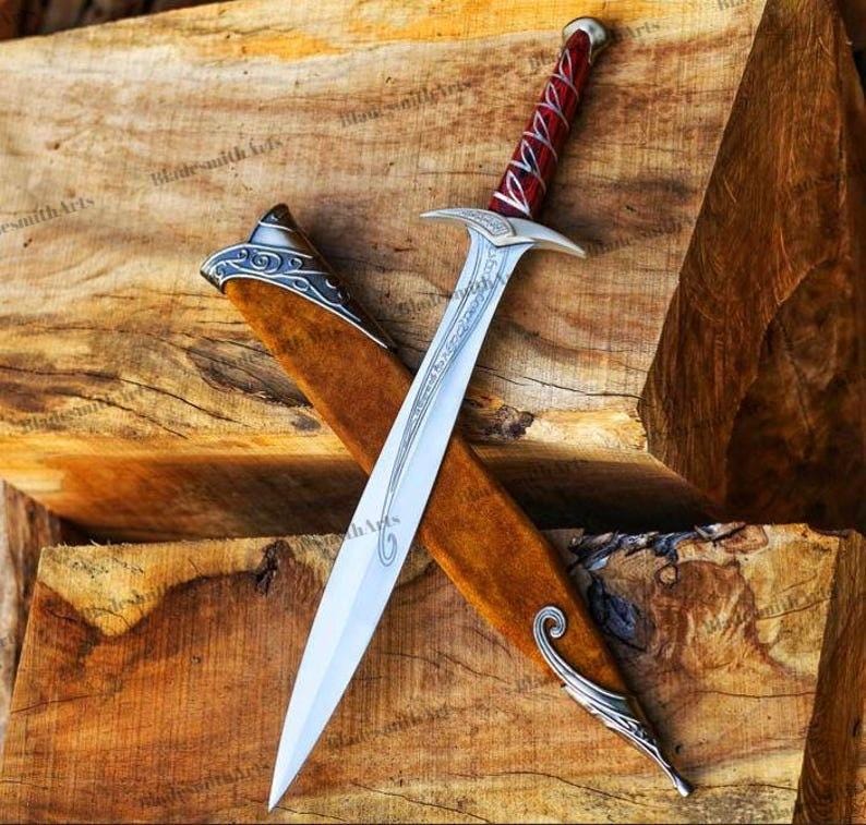 May include: A silver-toned metal sword with a pointed blade featuring etched line details. The handle is wrapped in a deep red material with silver accents. It is positioned diagonally across a brown wooden scabbard with silver metal trim.