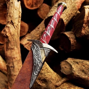 May include: A dagger featuring a red wood-grain handle with silver-colored metal runes, a silver-colored metal crossguard with intricate scrollwork engravings, and a brown leather-textured sheath partially covering the blade.