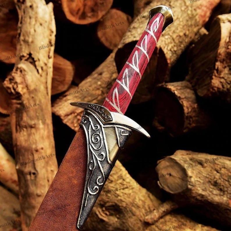 May include: A dagger featuring a red wood-grain handle with silver-colored metal runes, a silver-colored metal crossguard with intricate scrollwork engravings, and a brown leather-textured sheath partially covering the blade.