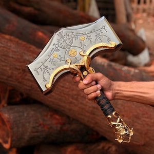 Pu&ograve; includere: Un grande martello in metallo con una testa color argento incisa con intricati motivi runici neri e accenti dorati. L'impugnatura &egrave; avvolta in materiale scuro con dettagli in metallo dorato alla base e vicino alla testa del martello.